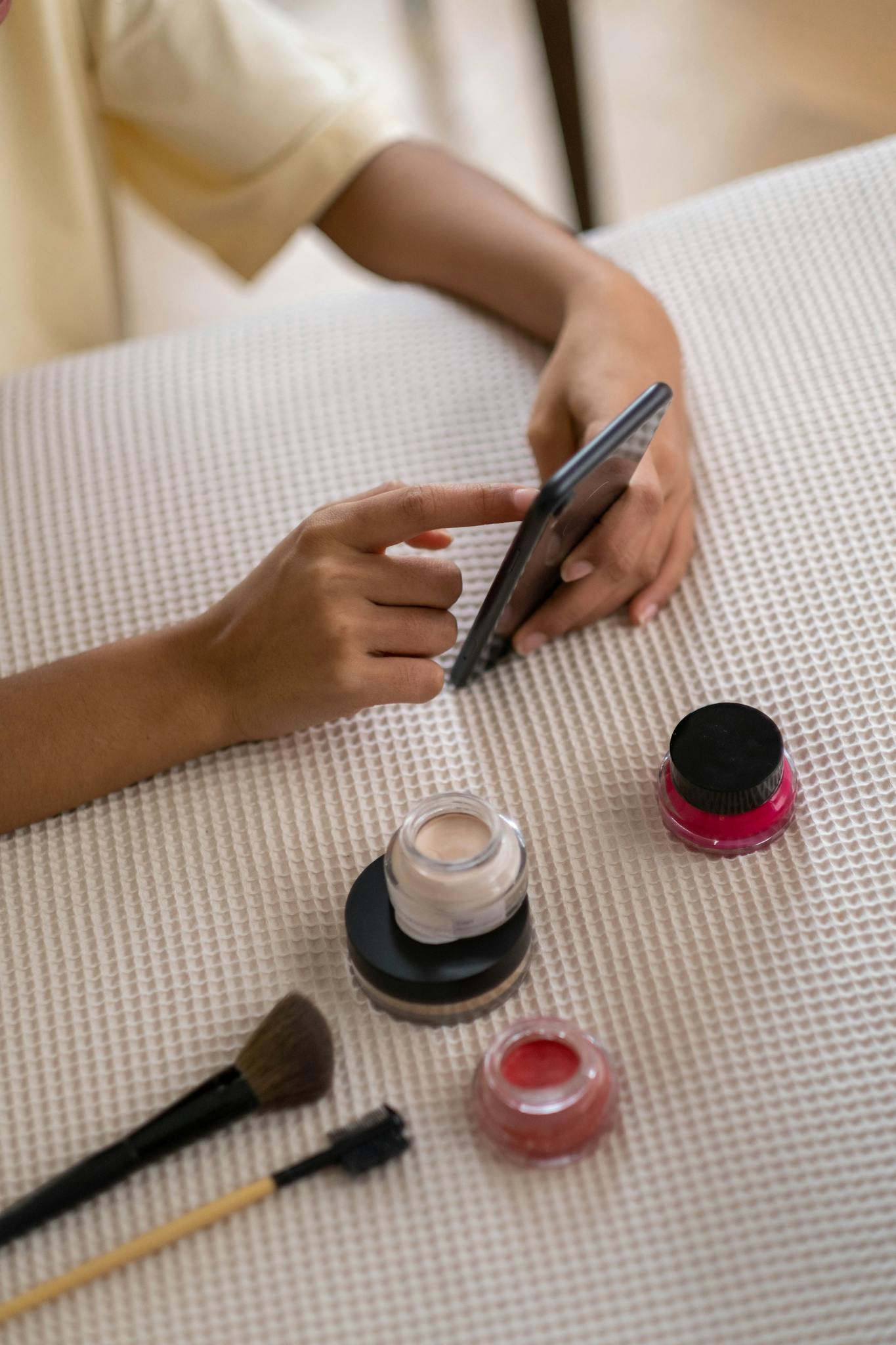 Close-up of a woman using a smartphone surrounded by beauty products and brushes, perfect for self-care themes.